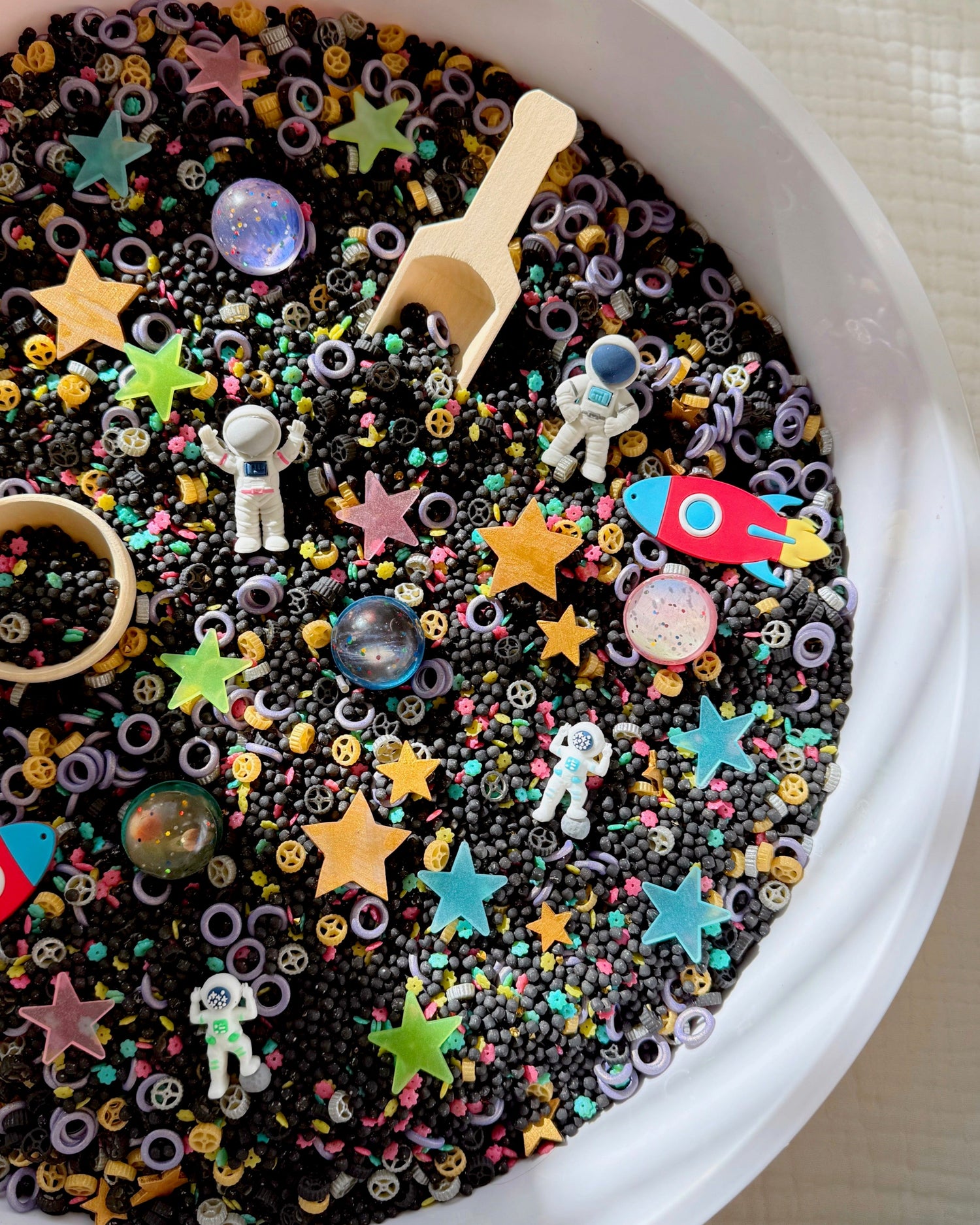 Outer space sensory bin with black couscous filler, astronaut figures, red rocket, planets and colorful stars