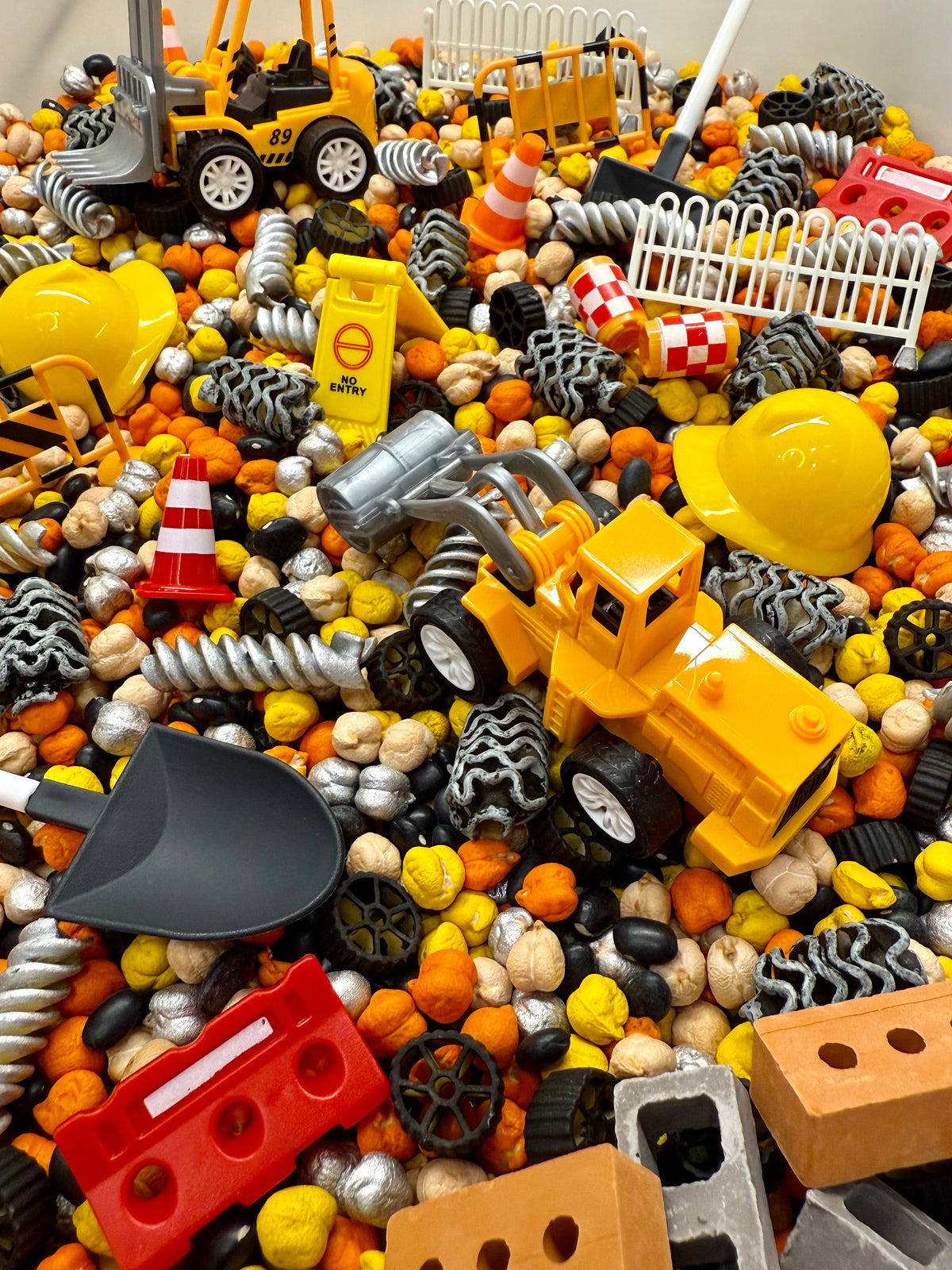 Construction sensory bin close-up with toy diggers, hard hats, bricks, fences and orange, yellow and black filler