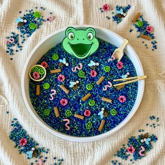 Lily Pad Lagoon sensory bin with frog face, blue filler, flies, worms and wooden tools