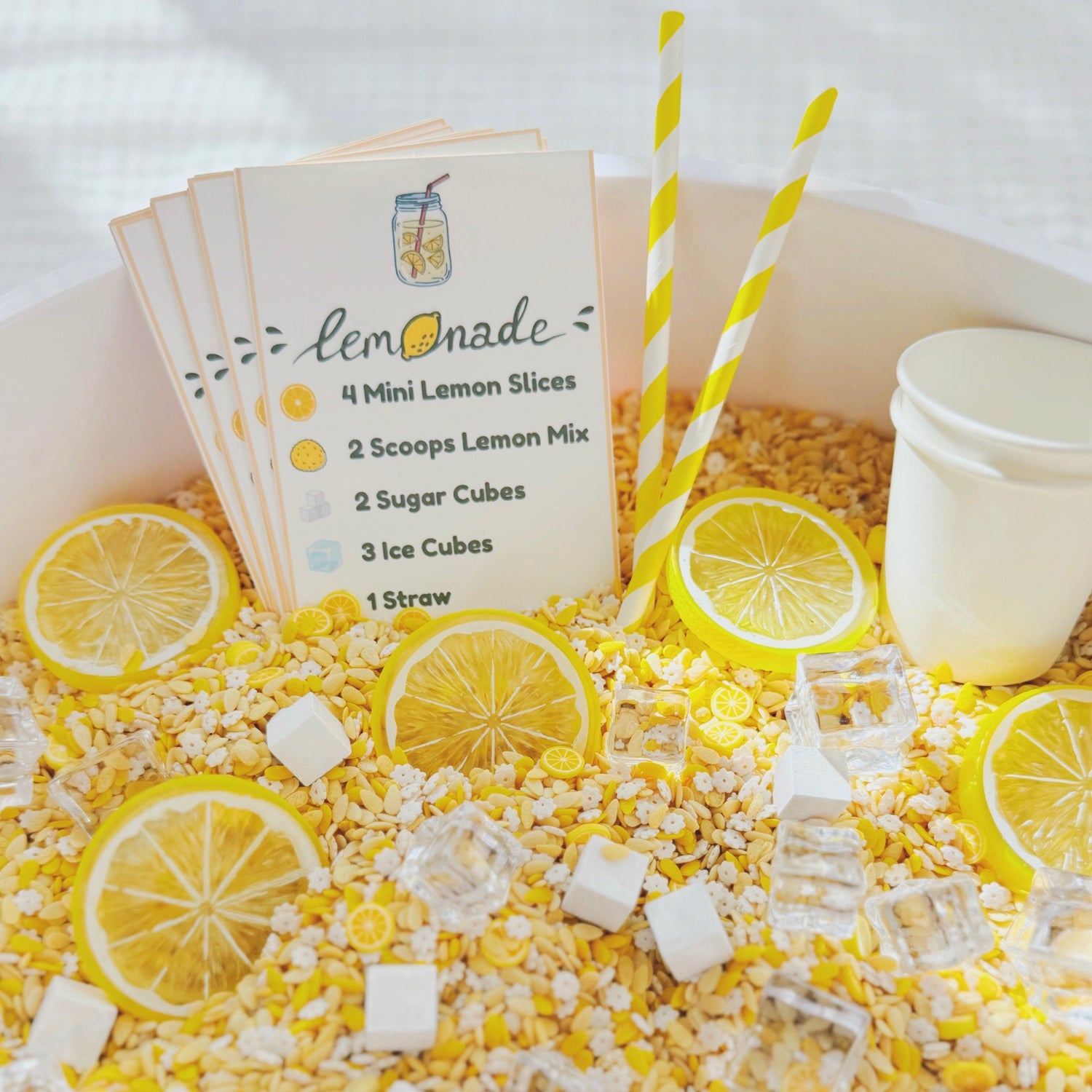 Lemonade stand sensory bin with yellow lemonade-themed filler, recipe cards, striped straws, lemon slices and sugar cubes
