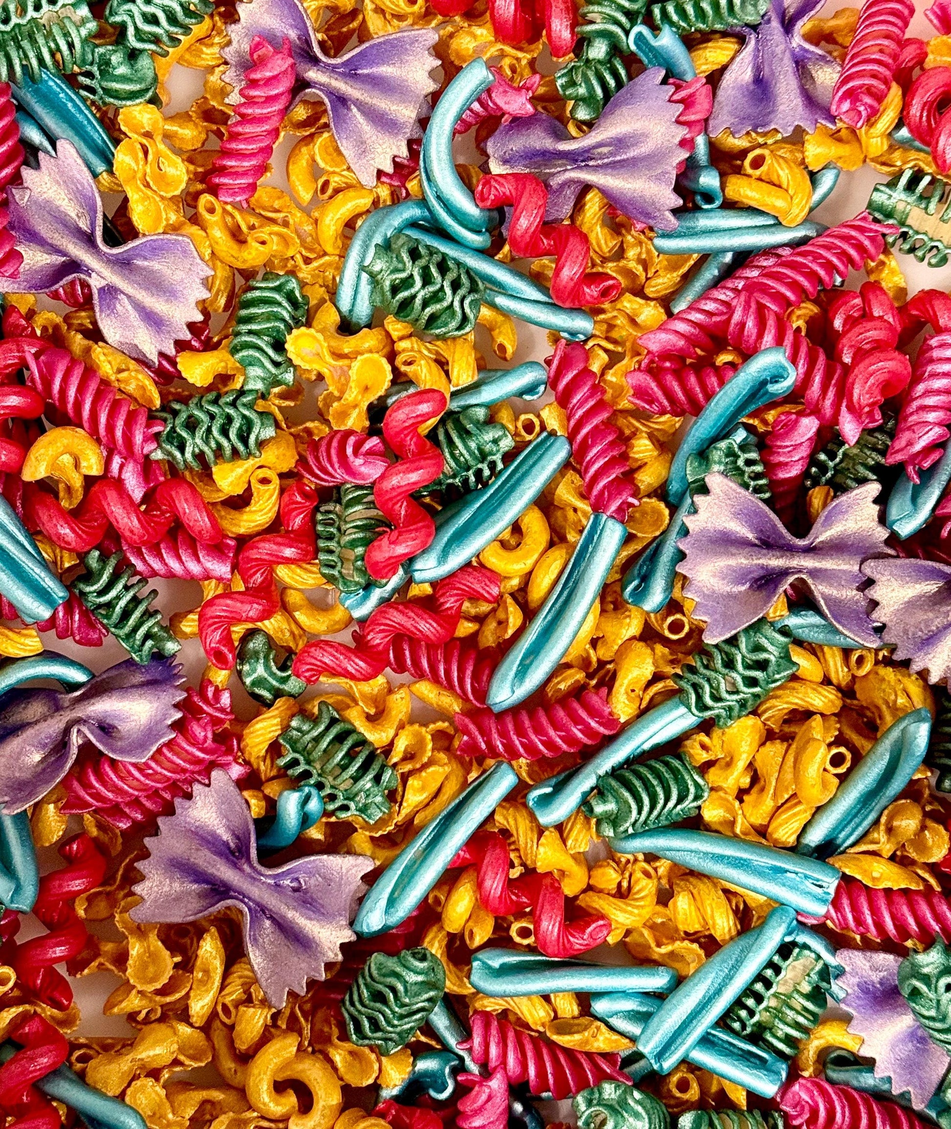 A colorful assortment of pasta shapes to be used as sensory bin filler. Pasta shapes include fusilli, farfalle, and elbow macaroni. The pasta is dyed in vibrant hues of red, yellow, teal, green, and purple. The image is a close-up, showcasing the variety of shapes and colors.